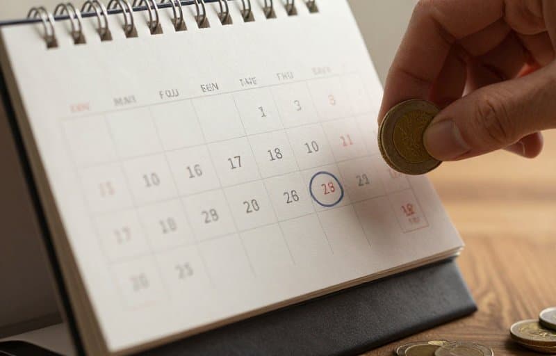 A monthly desk calendar with one date circled and a small stack of coins being lifted off the page, illustrating refund money leaving a later payout than the sale