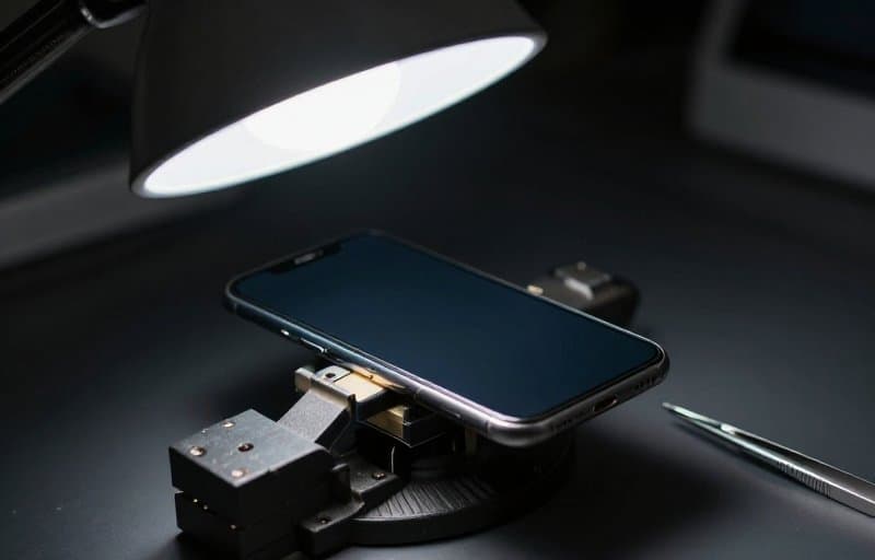 A smartphone held in a small bench vise under a work lamp with tweezers beside it, a device under test standing in for rehearsing a refund before it is real